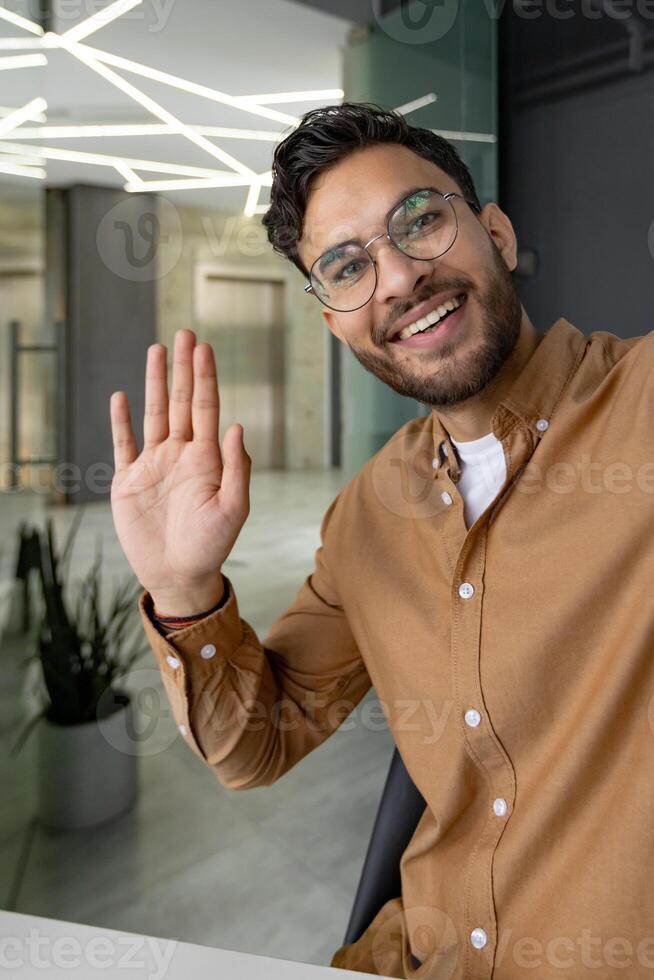 A happy, smiling man waves during a call in a modern office. The image captures friendly interaction and professional communication. photo
