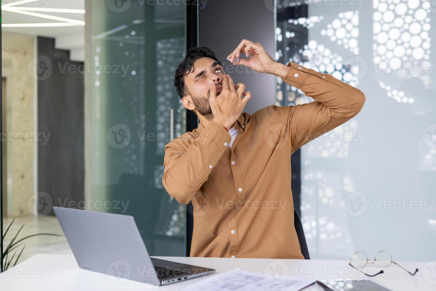 Man using eye drops while working at the office with laptop. Workplace discomfort and eye care. photo