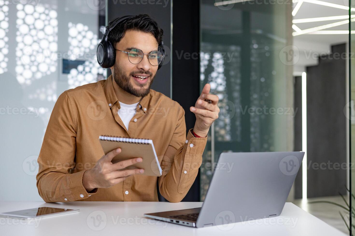 Man wearing headphones attending an online webinar while taking notes. He is sitting at a modern office desk with a laptop and notebook. photo
