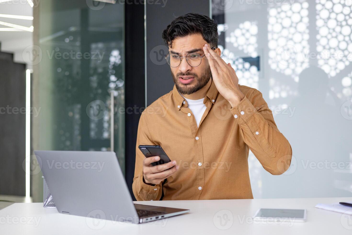 Frustrated man using phone and laptop in office, solving a problem. Concept of technical issues, business stress, and workplace challenges. photo