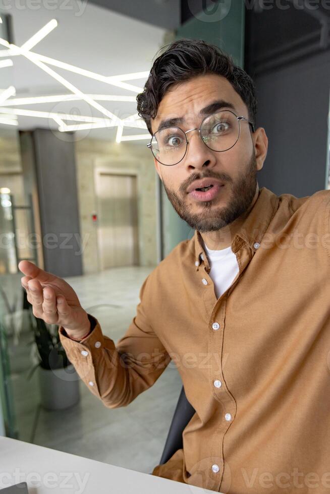Vertical close-up photo of a worried and serious young Muslim man in a shirt and glasses sitting at a desk in the office and talking on a call, holding a phone in his hand.