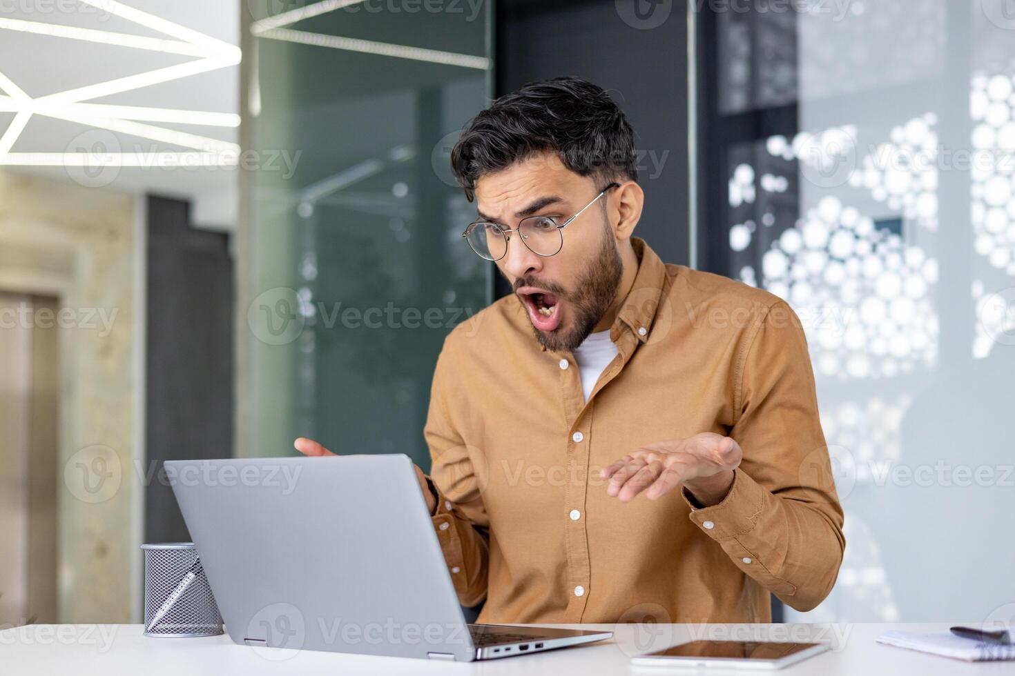 Young man in glasses expressing shock while looking at his laptop in a contemporary office setting photo