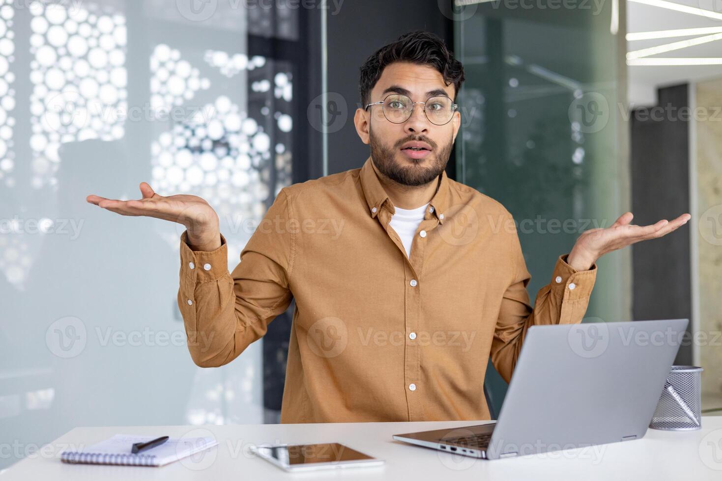 Young man with glasses in an office environment sitting in front of a laptop, expressing confusion and uncertainty with open palms and shrugging gesture. photo