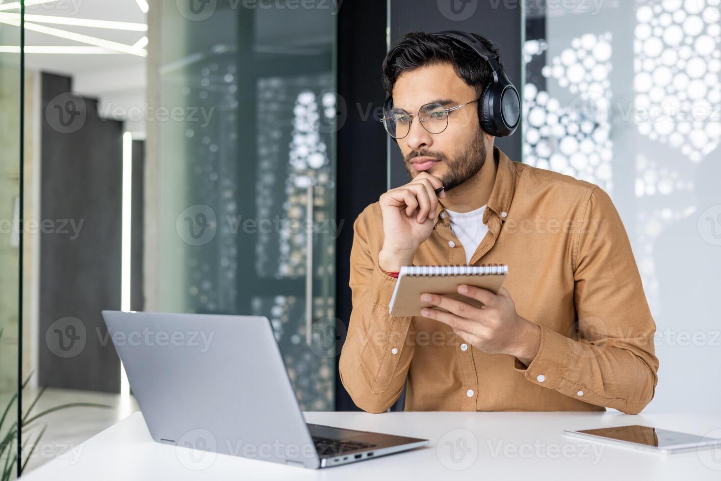 Thinking concentrated man studying sitting inside office at workplace, businessman watching training course, jotting down data in notebook, using laptop, employee upgrading skills, call. photo