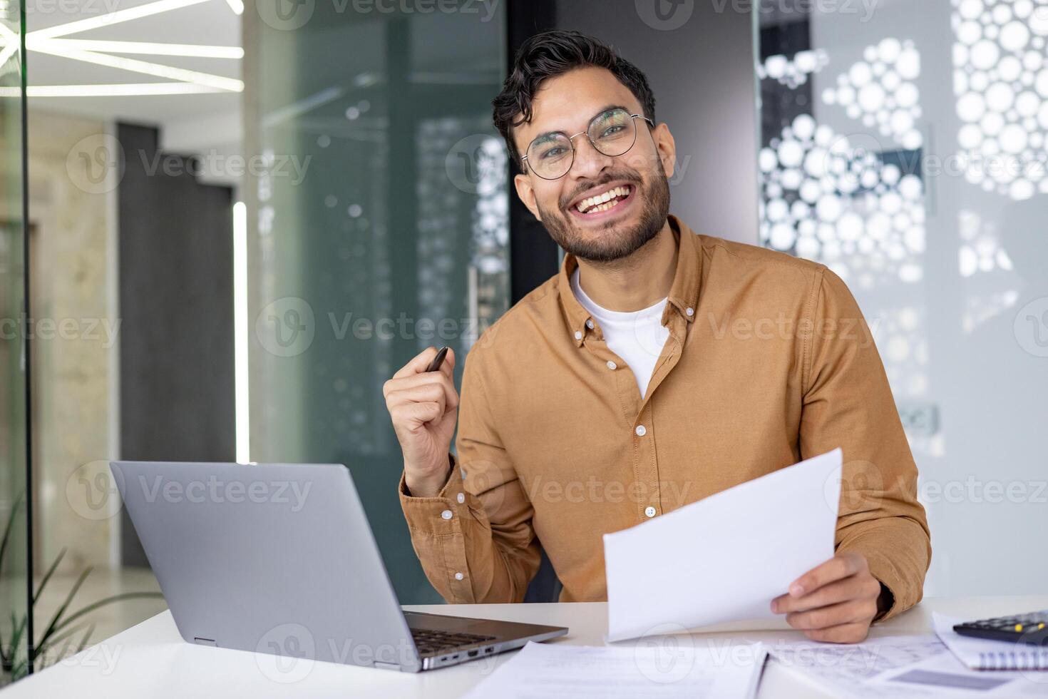 Happy young professional working in a modern office space with a laptop and documents, showcasing success and productivity. photo