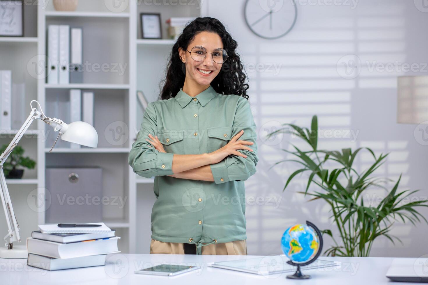 Confident female teacher smiling at her desk in a modern classroom setting with books, a lamp, and a globe photo
