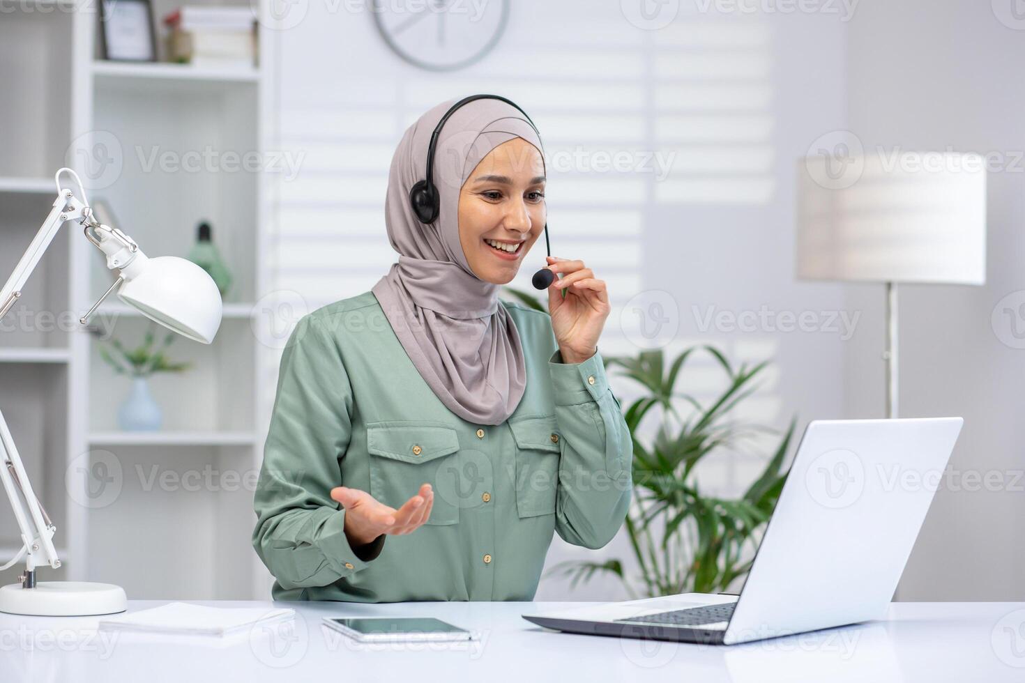 Smiling woman in a hijab using a headset on a call, working on a laptop in a modern home office. photo