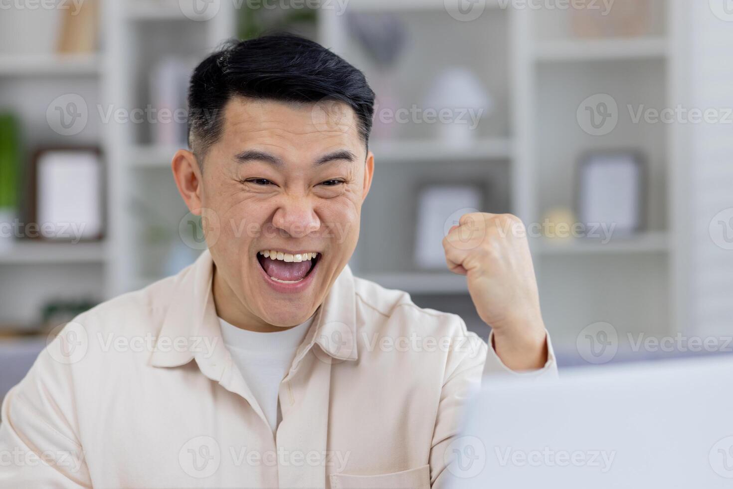 Joyful middle-aged Asian man in a casual shirt celebrating a triumph in front of his laptop, expressing excitement and happiness in a home office setting. photo