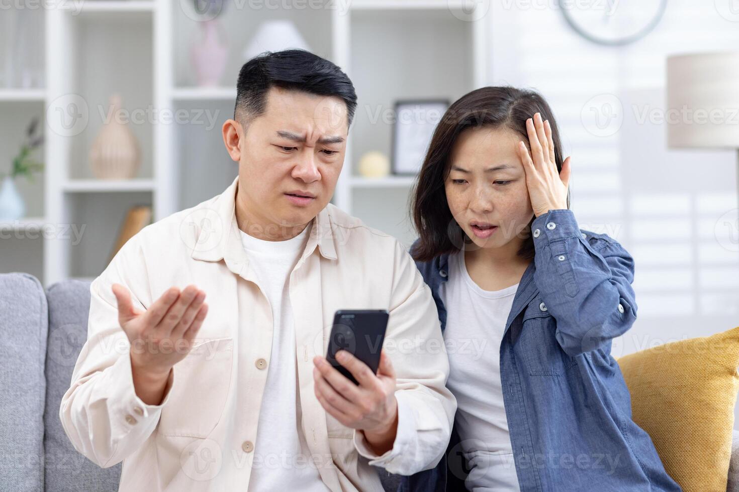 Concerned couple reacting to something on a smartphone screen, showing surprised and confused expressions, sitting in a living room photo