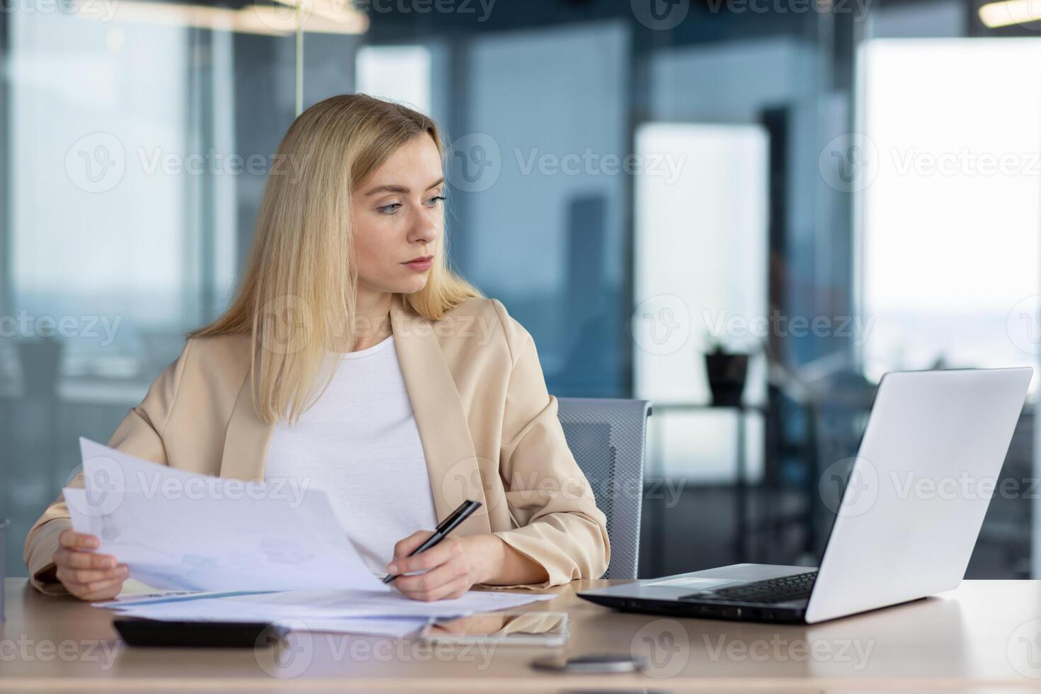 Professional businesswoman concentrating on work, analyzing documents while using a laptop at her desk in a contemporary office setting. photo