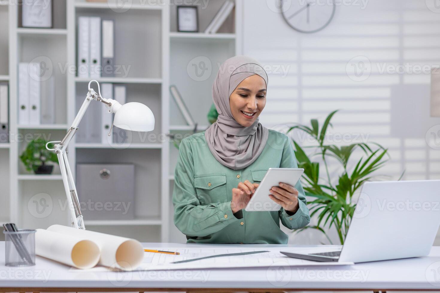 Confident female architect wearing hijab working on tablet at her desk in a modern office environment. Professional and focused on her architectural projects. photo