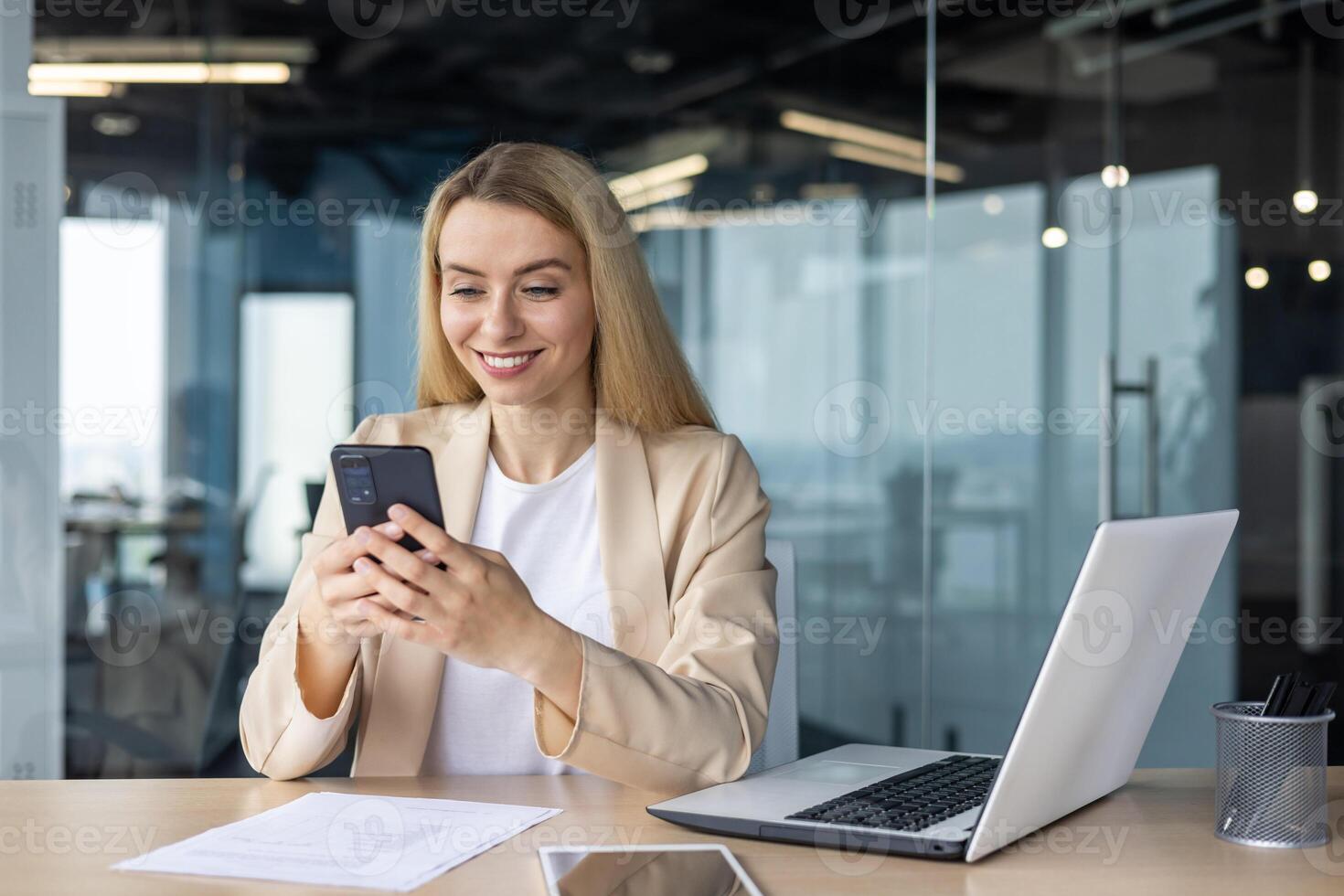 exitoso hermosa y contento mujer de negocios trabajando dentro oficina a lugar de trabajo, rubia participación teléfono utilizando en línea solicitud, sonriente sentado a mesa con computadora portátil, satisfecho con trabajo logro. foto