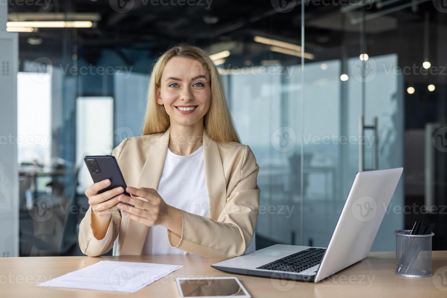 Portrait of a young businesswoman sitting in the office at a desk, using a mobile phone and smiling at the camera. photo