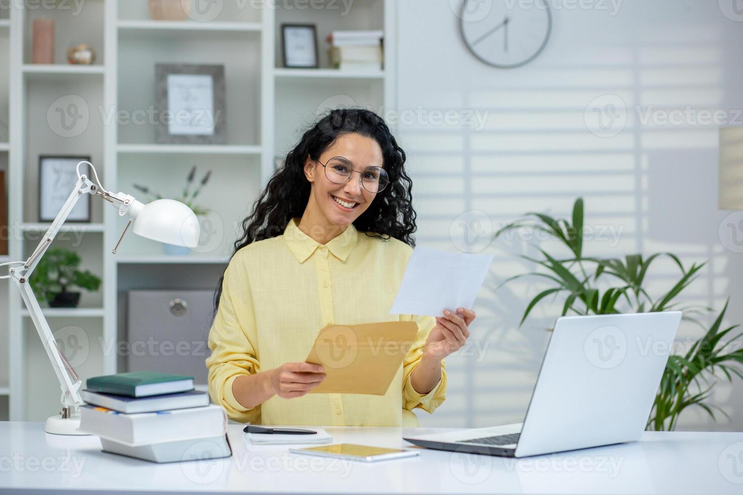 Smiling woman holding an envelope and letter while standing in a bright home office. Happy moment as she reads good news or a positive notification. photo