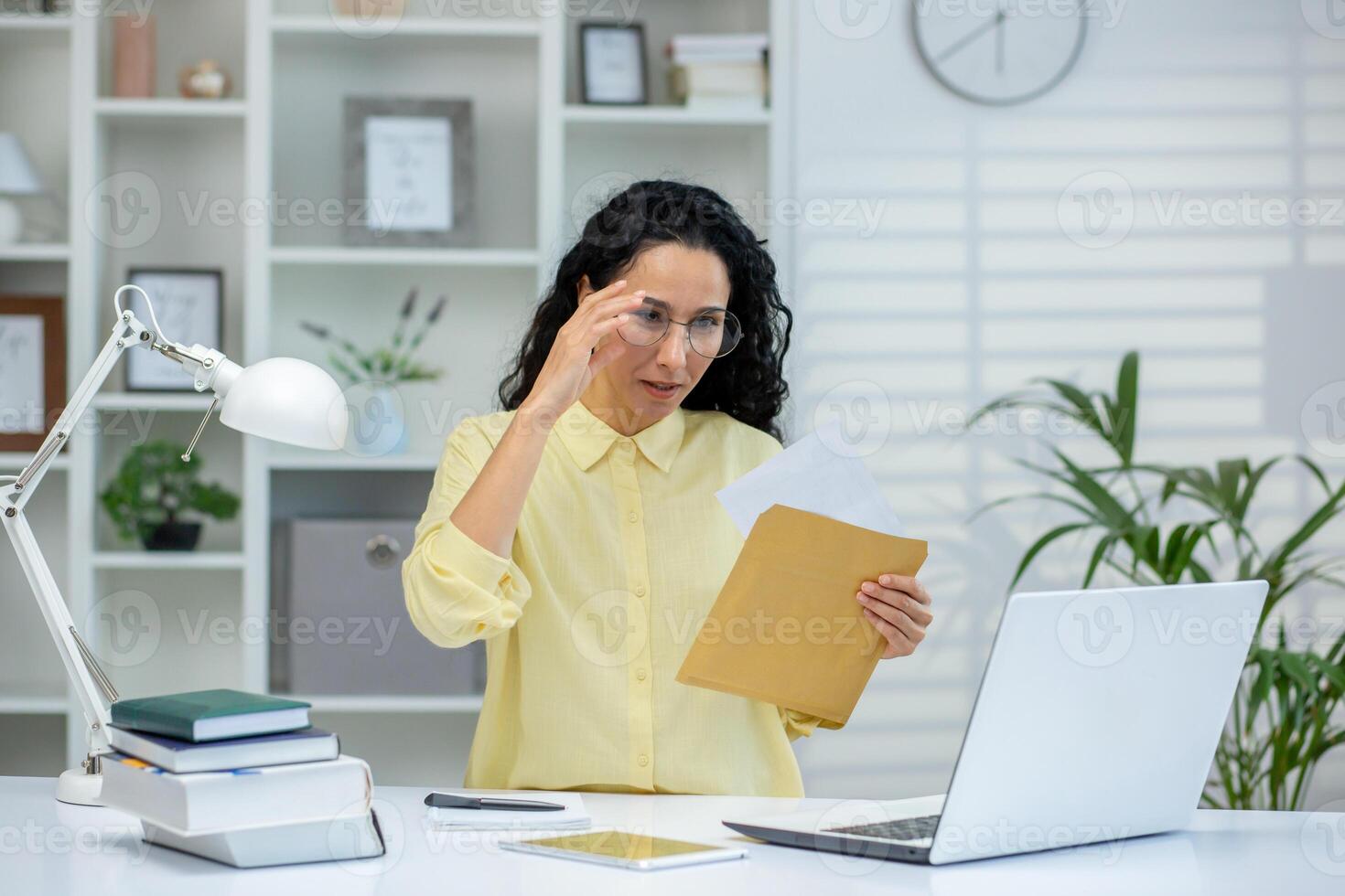 Woman working from home office, reviewing documents and reading contents of an envelope. Working on a laptop with a focused expression. photo