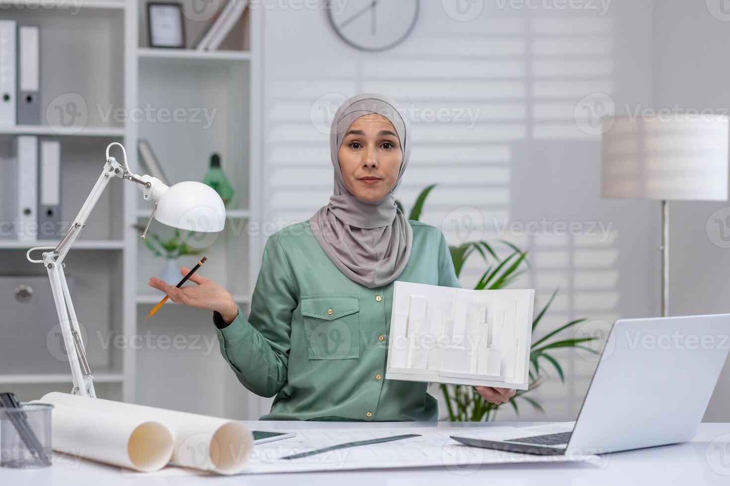 A confused female architect in a modern office holding a city model while sitting at a desk with architectural drawings and a laptop. photo