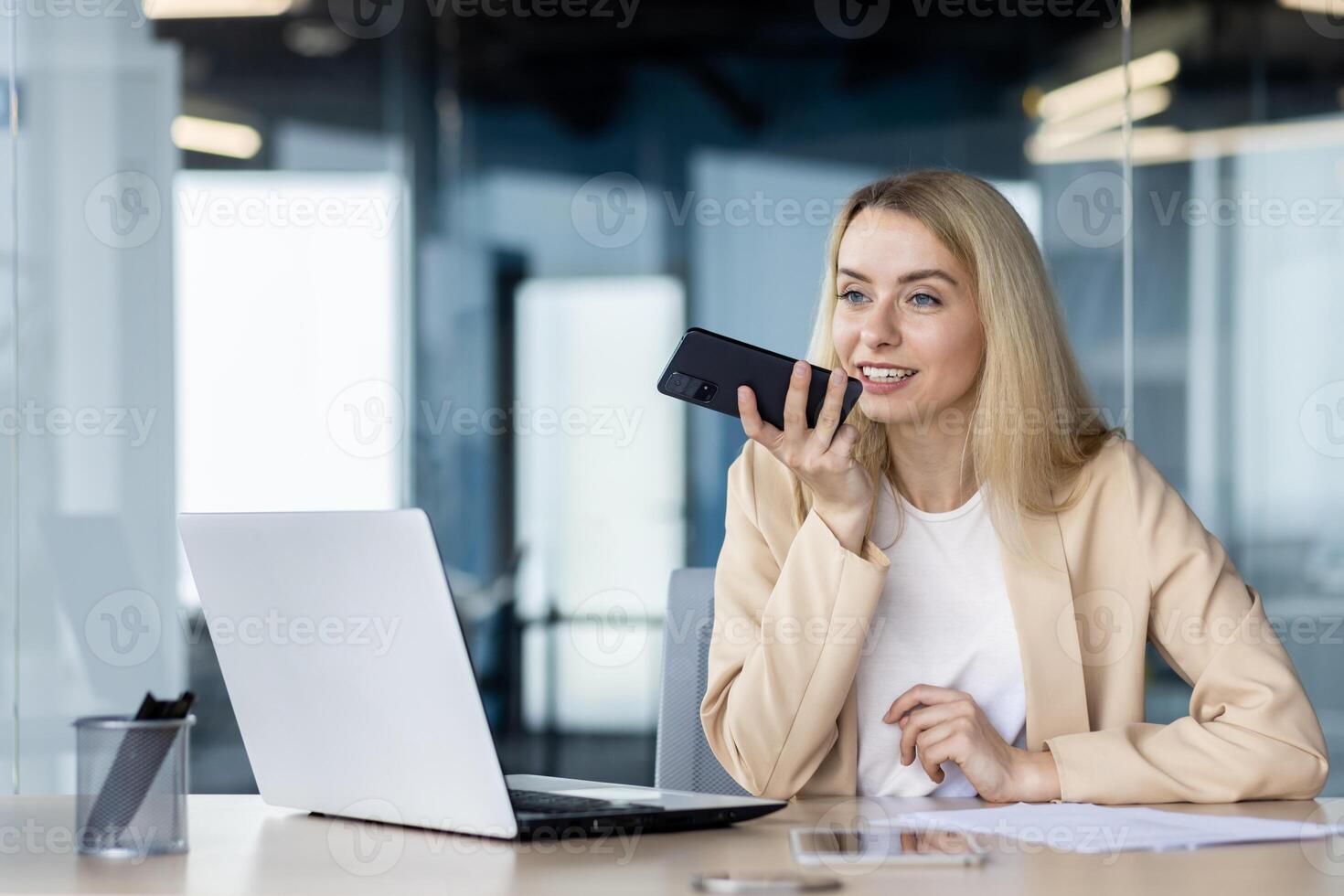 Confident businesswoman holding phone for a voice message in a contemporary workspace with a laptop and documents. photo
