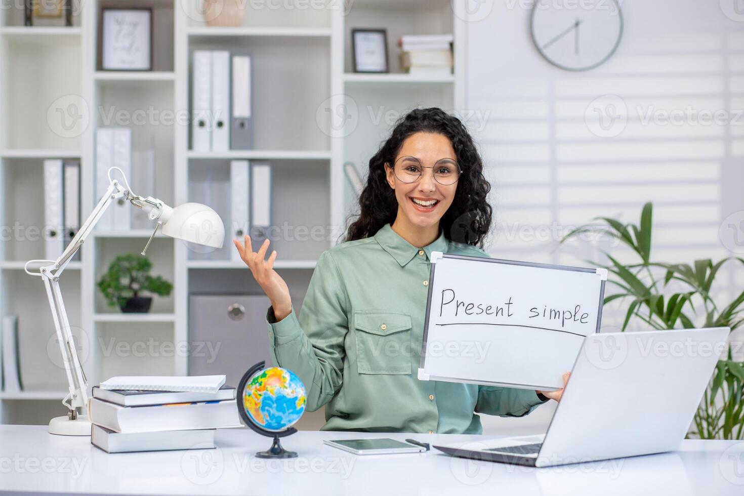 Smiling female teacher holding whiteboard with 'Present simple' text, teaching English online from a modern office setup. photo