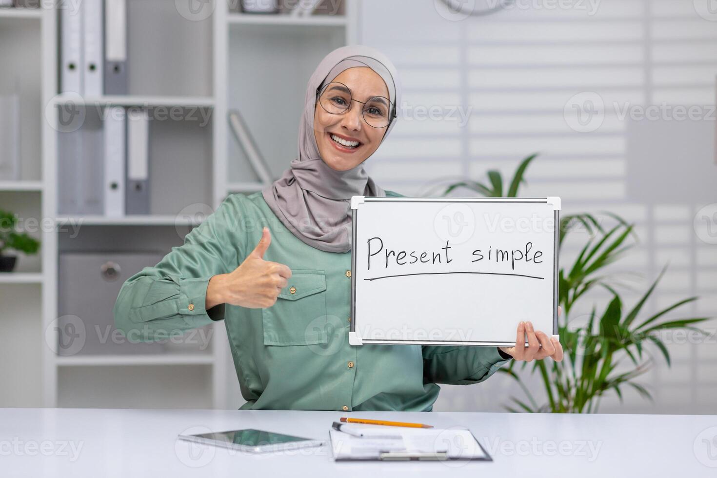 Happy teacher holding a whiteboard with present simple written on it and giving a thumbs up in a classroom setting. photo