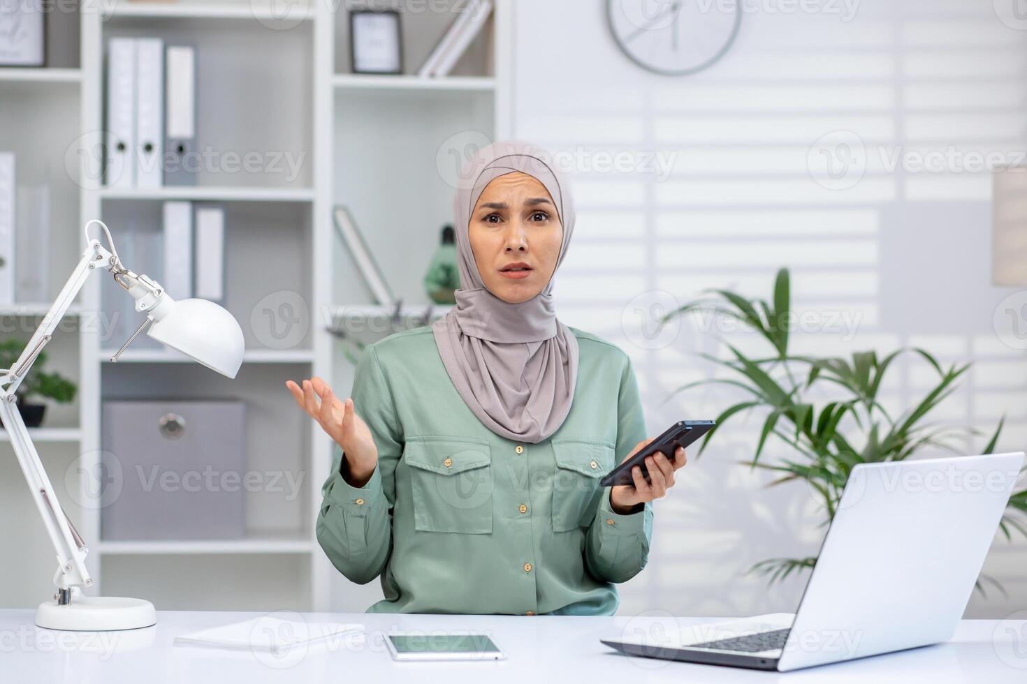 Woman in hijab working from home office, holding smartphone and gesturing in confusion. Modern workspace with laptop and documents. photo