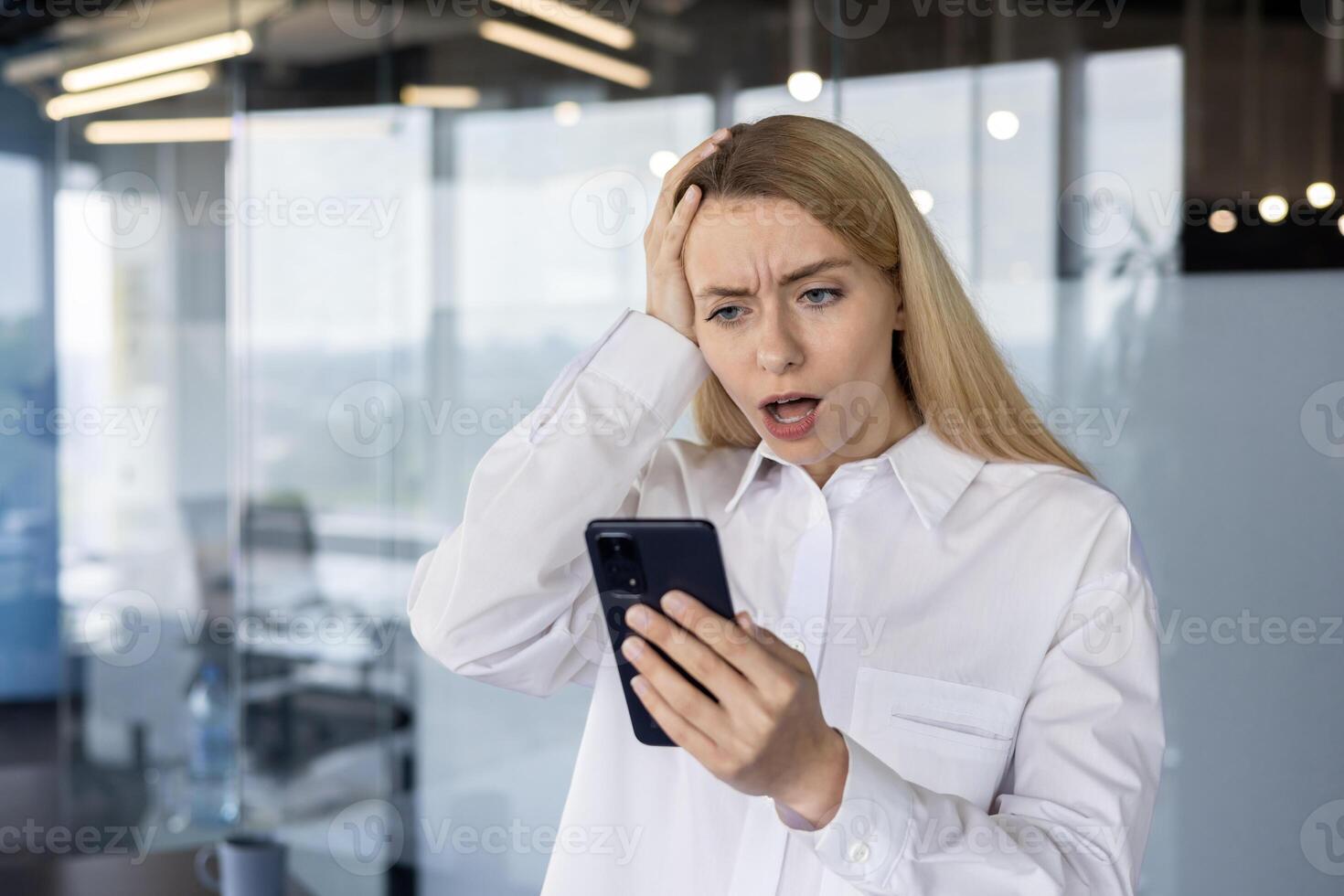 Stressed young businesswoman in a modern office, reacting to a surprising text message on her smartphone with a concerned expression. photo
