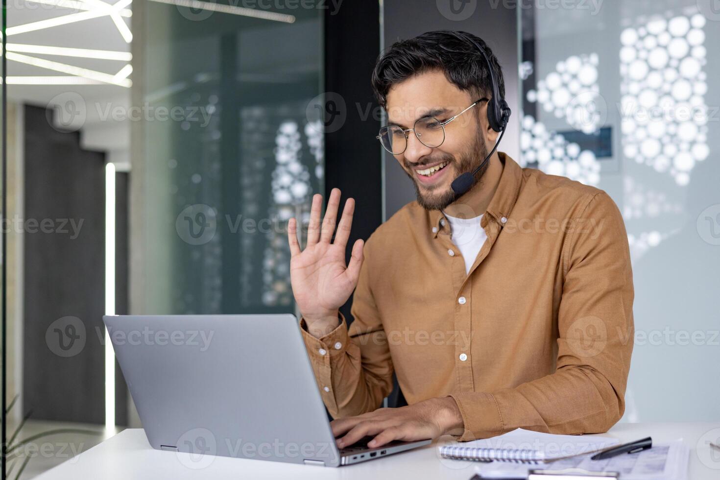 A smiling man wearing a headset and waving during an online call in a modern office setting. photo
