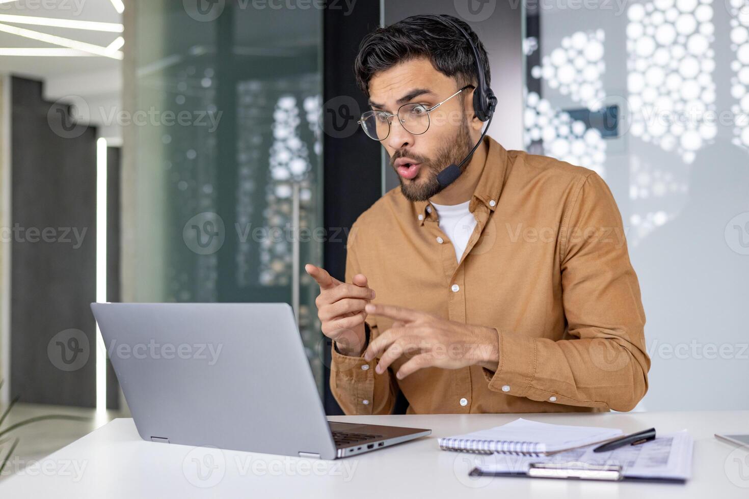 A serious young Indian man sits in the office at the desk in a headset and looks shocked at the laptop screen, reads the news, talks on a call. photo