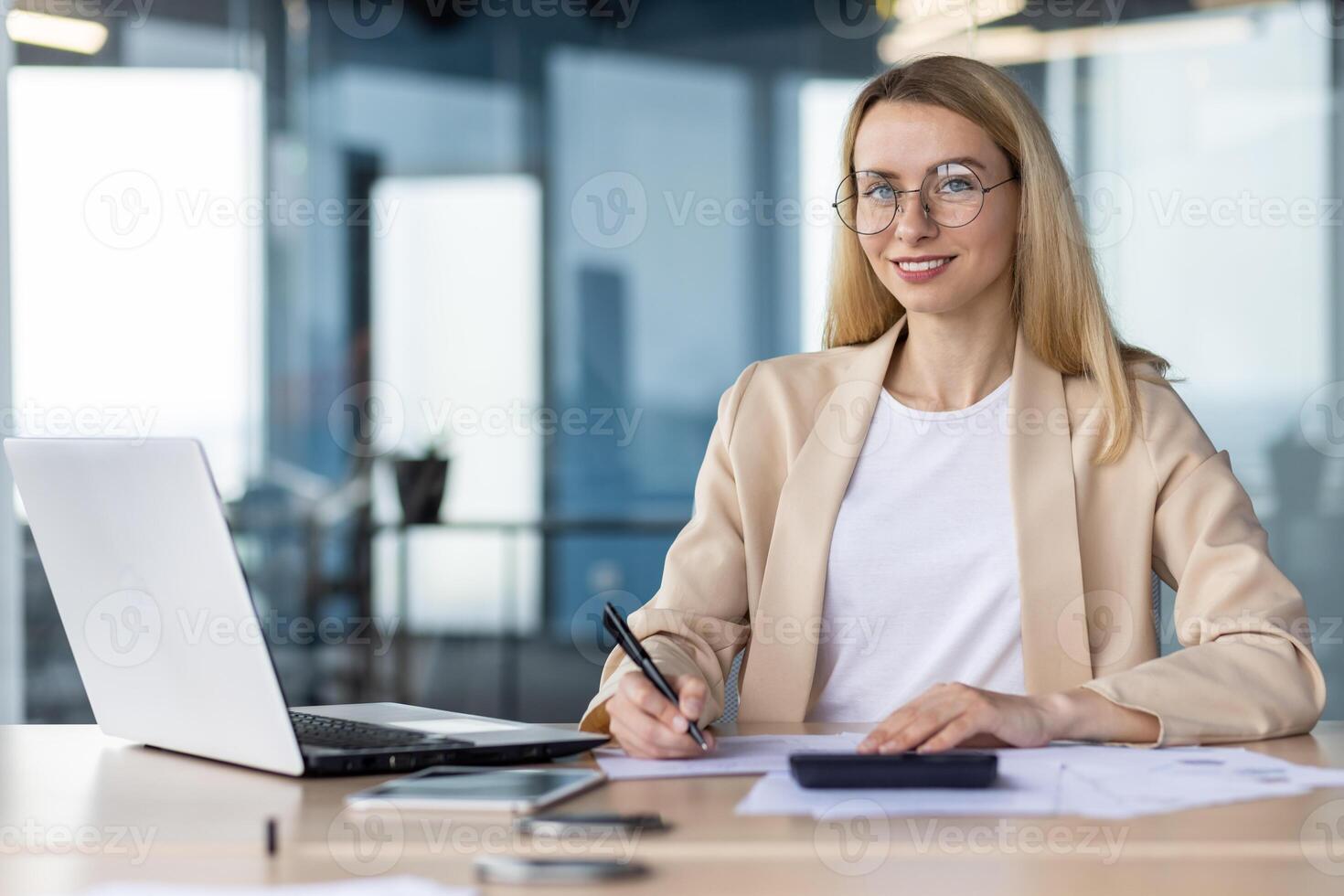 Portrait of a young female student, intern. Sitting in the office at the table, studying online from a laptop, looking at the camera with a smile. photo