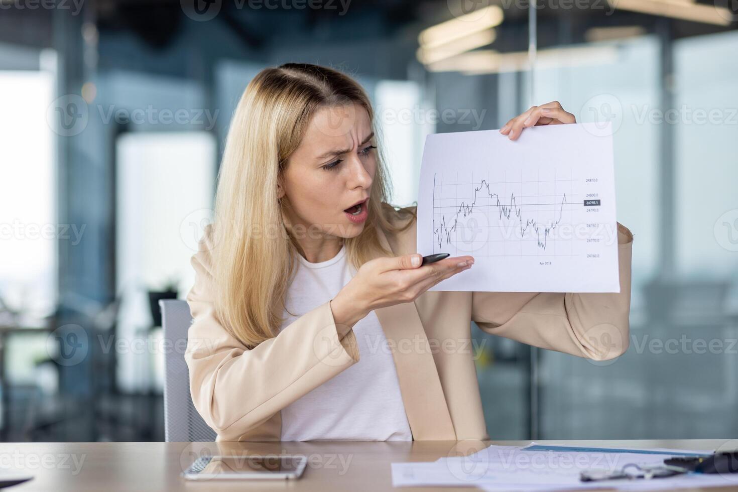 Angry and worried young business woman working in office with documents. Sits at the table in front of the camera and points with his hand to papers with graphs. photo