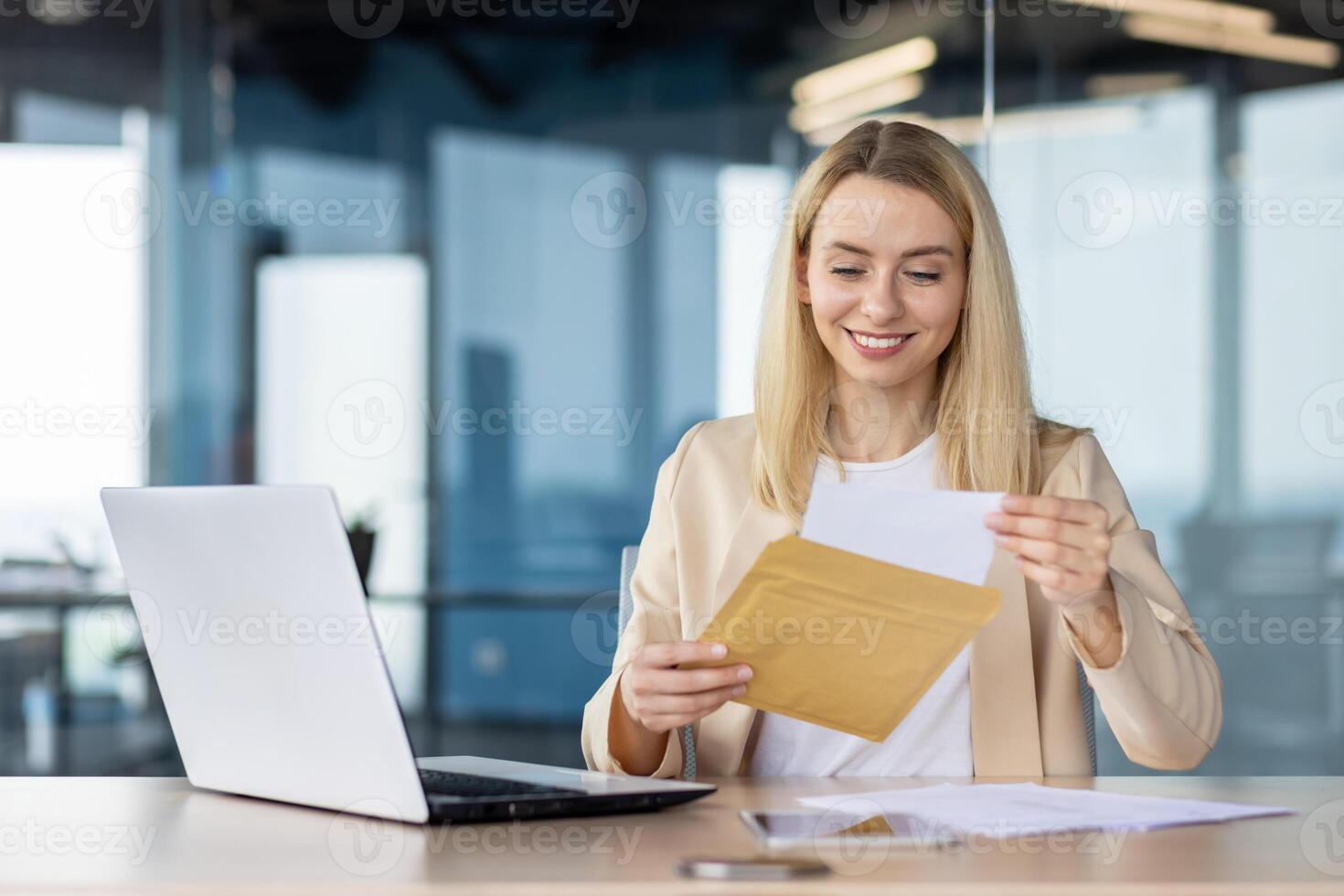 Professional woman in office attire smiling as she opens a yellow envelope, feeling excited and content at her desk with a laptop. photo