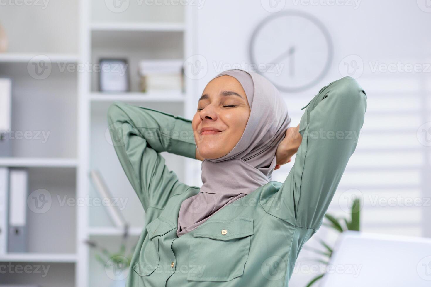 Woman in hijab stretching and relaxing at office workspace with a blissful expression. Concept of work-life balance, well-being, and relaxation photo