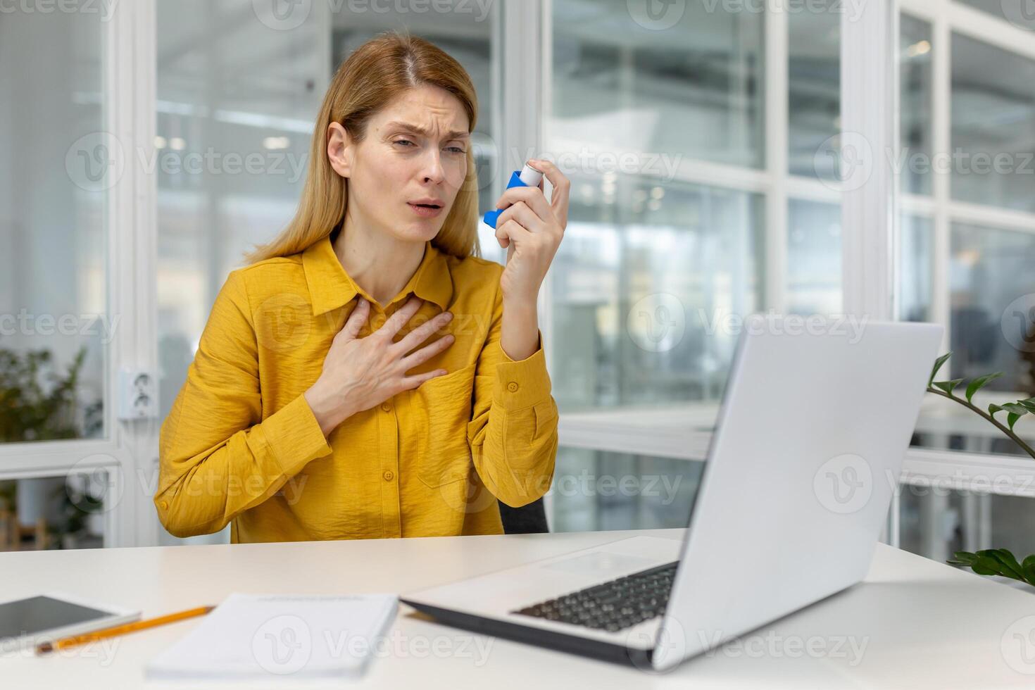 Woman experiencing asthma attack using inhaler while working on laptop in a modern office environment. Business stress and health issue concept. photo