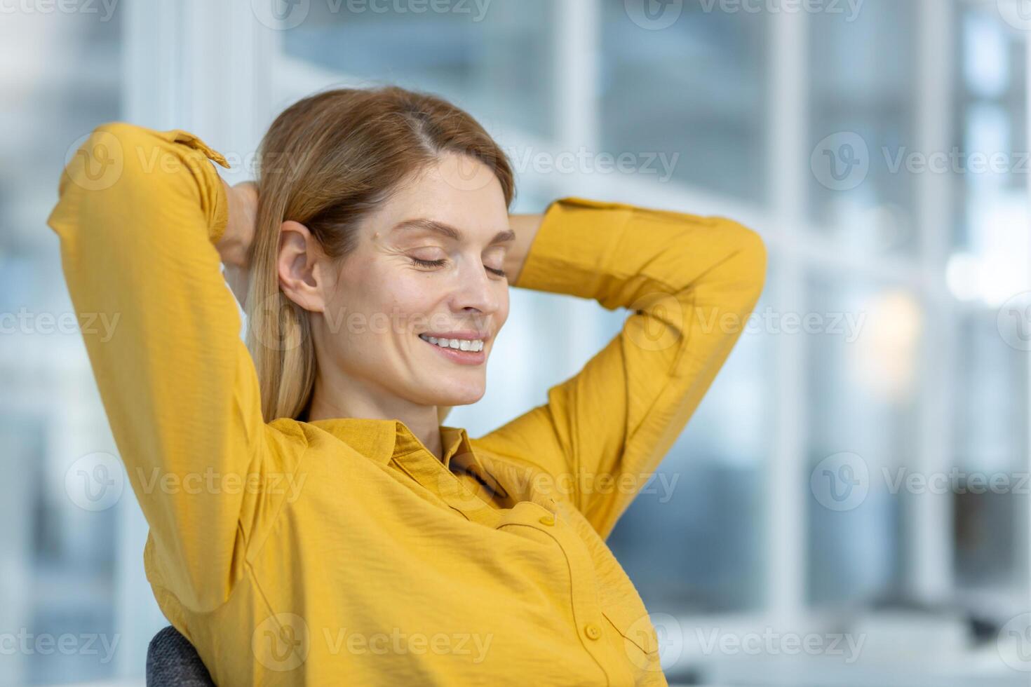 Woman wearing a yellow shirt relaxing with her eyes closed in an office environment. She is smiling and looking content. photo