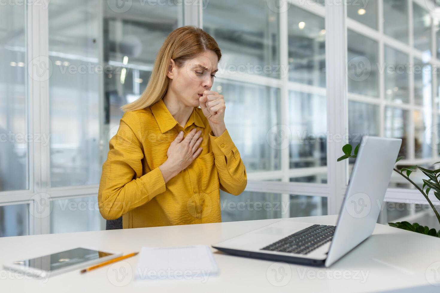 Woman experiencing a cough and chest pain while working on a laptop in a modern office environment. photo