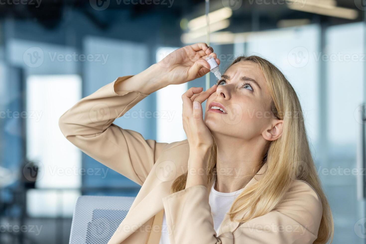 A businesswoman in a beige suit applies eye drops while looking upward, expressing discomfort, in a bright office environment. photo