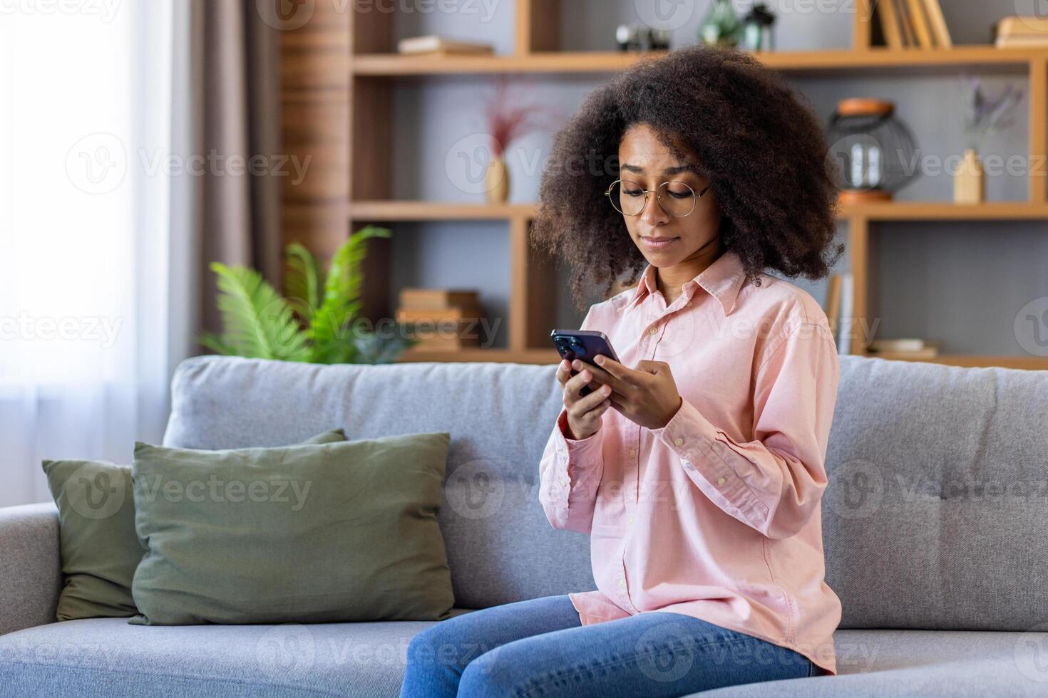 Serious thinking woman using phone sitting at home in living room, Hispanic woman with curly phone, typing text message, thinking, reading internet pages, browsing social networks. photo