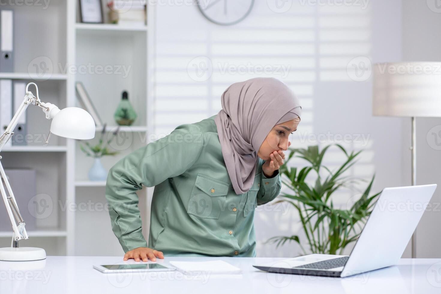 A woman in a hijab feeling sick and experiencing nausea while working at her desk in an office, gazing at her laptop. photo