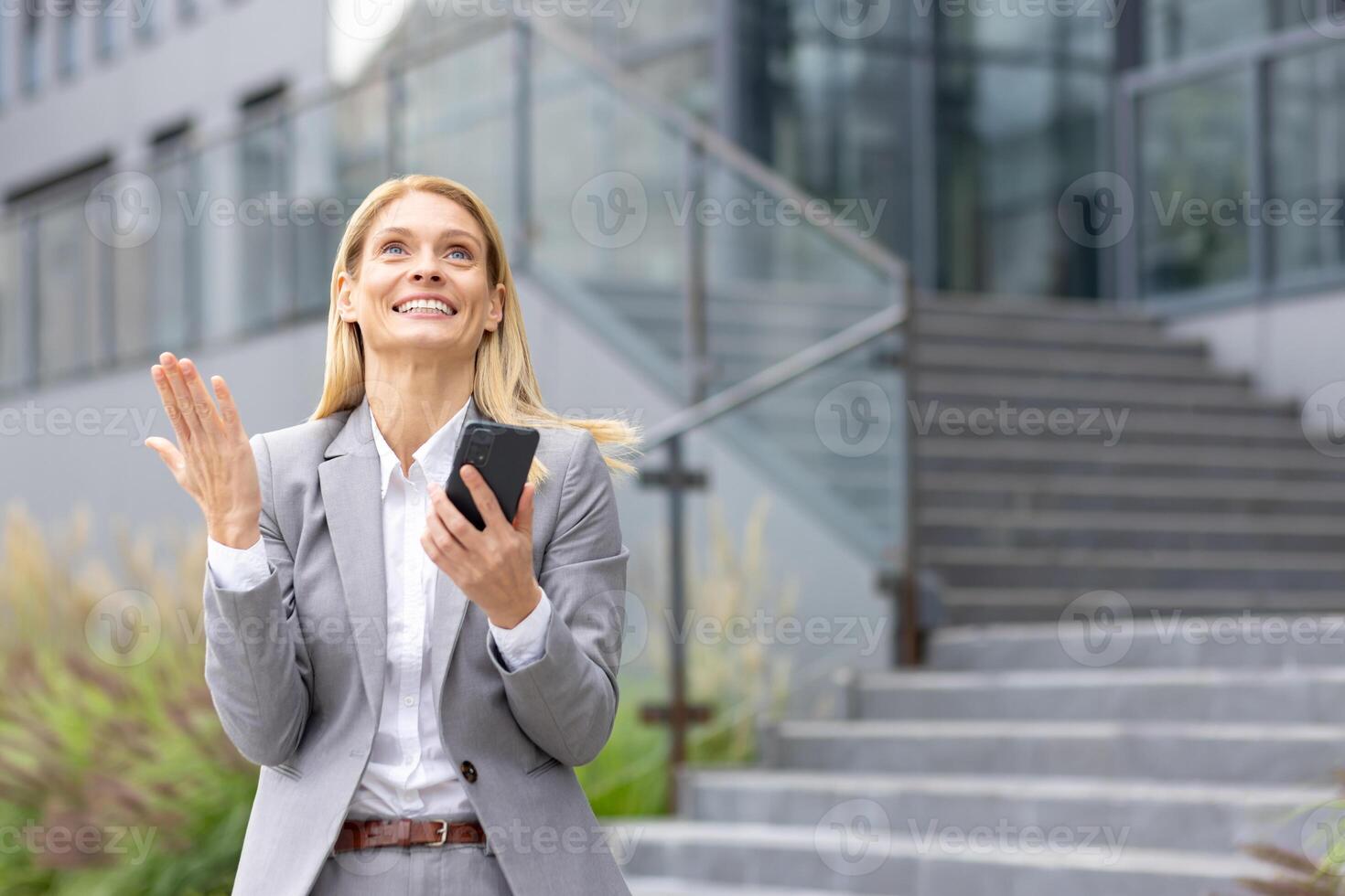 An accomplished businesswoman in a gray suit radiates happiness and selfassurance while celebrating outside a sleek office building, holding a mobile phone, standing on the steps photo