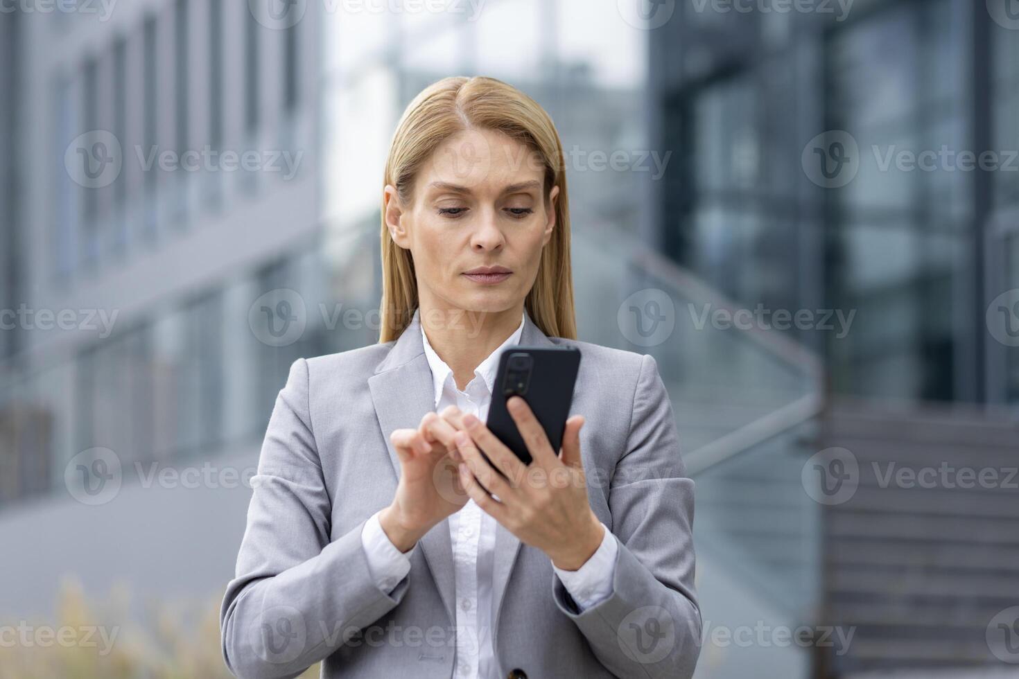 Focused businesswoman in gray suit browsing her smartphone against a modern office building backdrop. Captures a moment of technology use in a corporate environment. photo