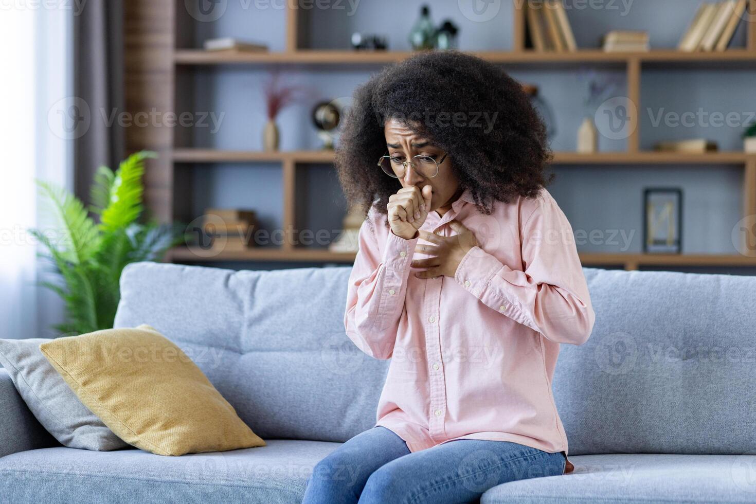 A middle-aged woman with glasses is coughing into her hand while sitting on a gray couch. The background features shelves with books and plants, creating a cozy living room atmosphere. photo