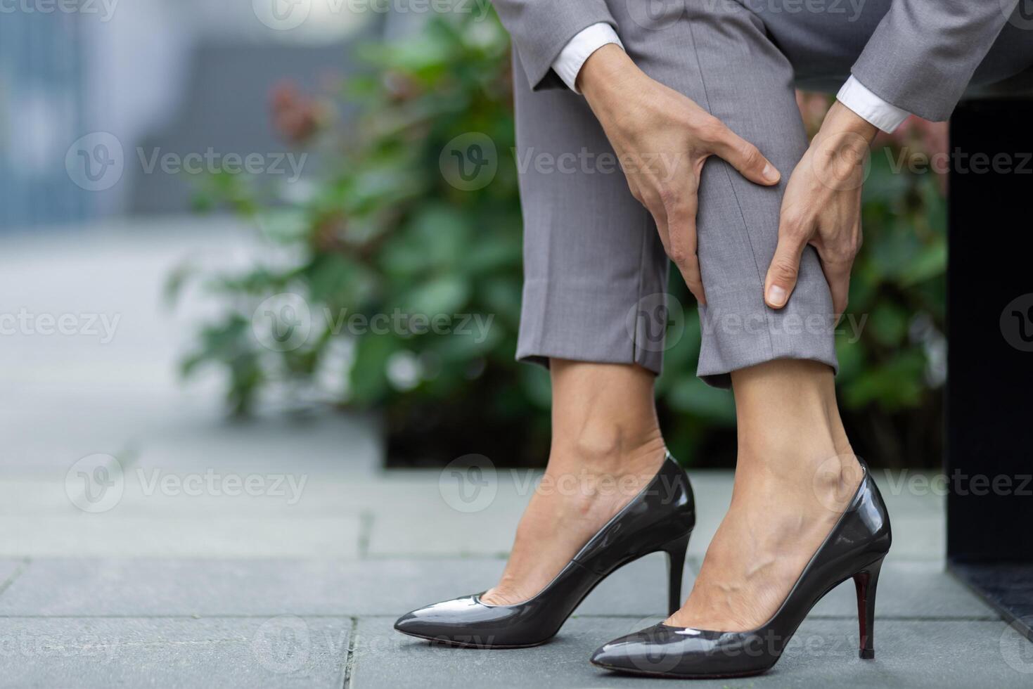 A close-up shot of a professional woman in a business suit massaging her calf on a city street, illustrating discomfort from wearing high heels. photo
