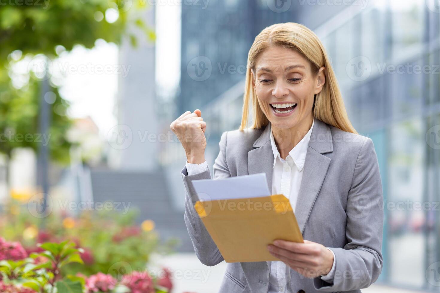 A joyful businesswoman, professionally dressed, celebrates outdoors in an office courtyard upon receiving positive news in an envelope. She reflects happiness and success in her expression photo