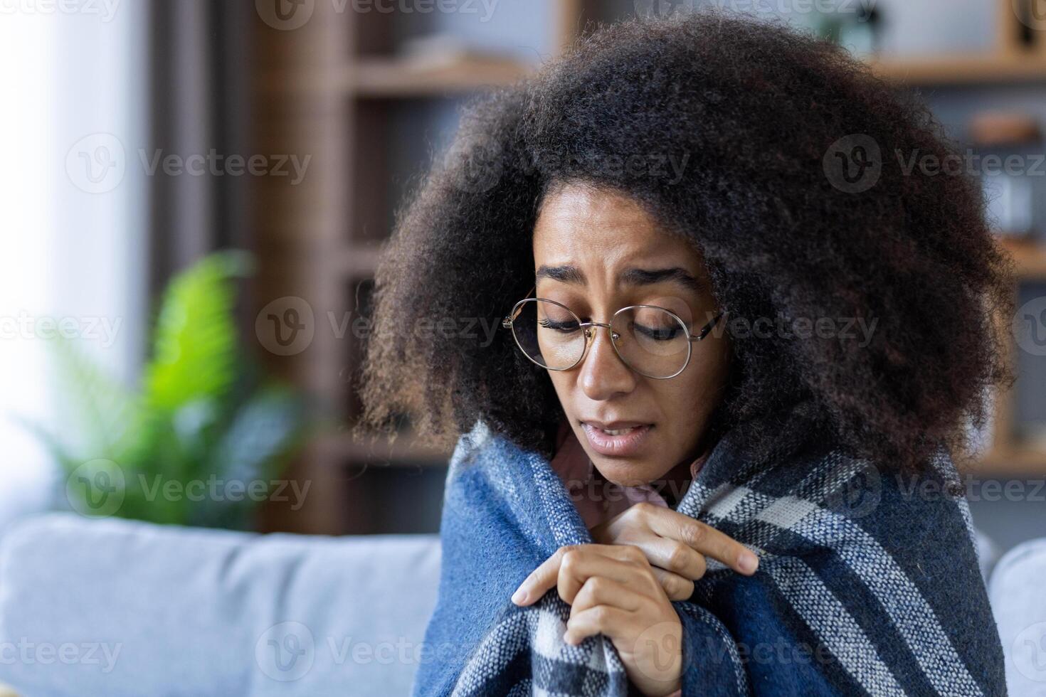 A young woman is wrapped in a blue plaid blanket, feeling cold and showing a sad expression. She has glasses and an afro hairstyle. Background includes a green plant and blurred shelves. photo