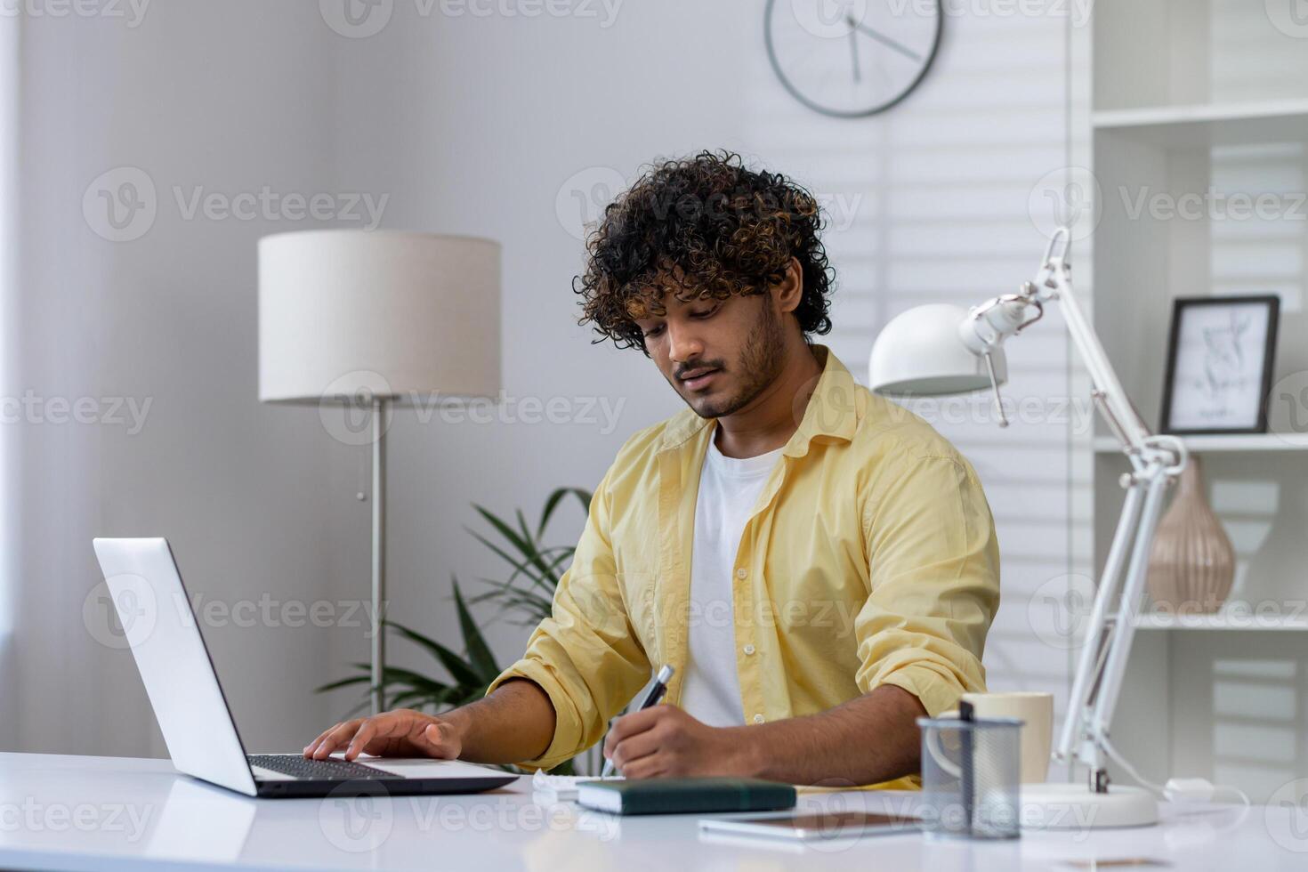 Man in a yellow shirt working remotely from home, using a laptop and taking notes in a notepad at a modern desk. photo