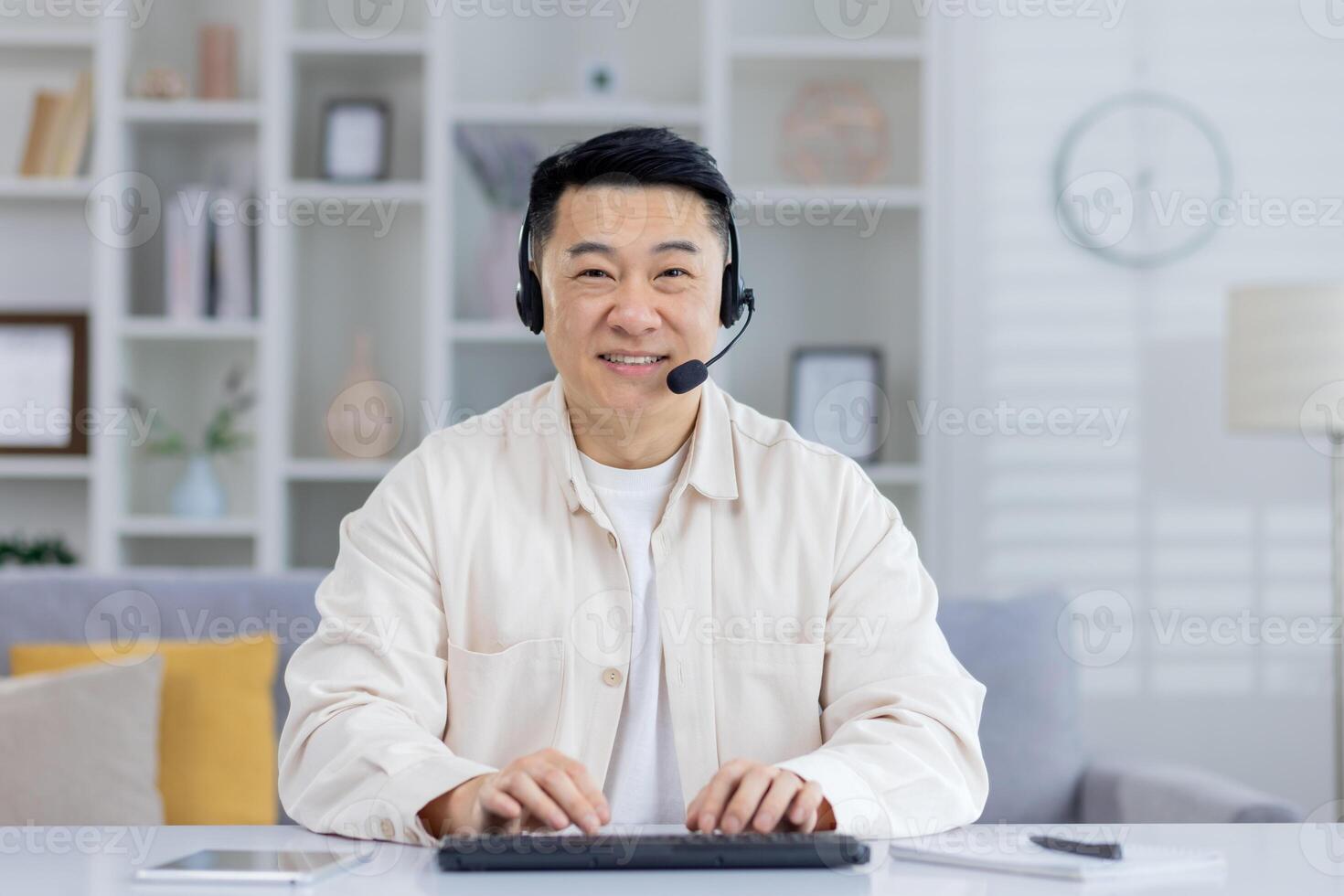 A middle-aged Asian man smiles warmly while using a headset and working on a keyboard in a bright, modern home office setting. photo