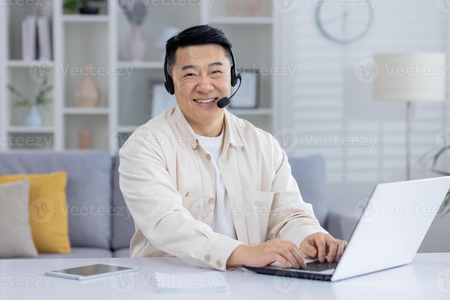 Happy middle-aged Asian man using a headset and laptop in a well-decorated home office. He exudes professionalism and contentment while working remotely. photo