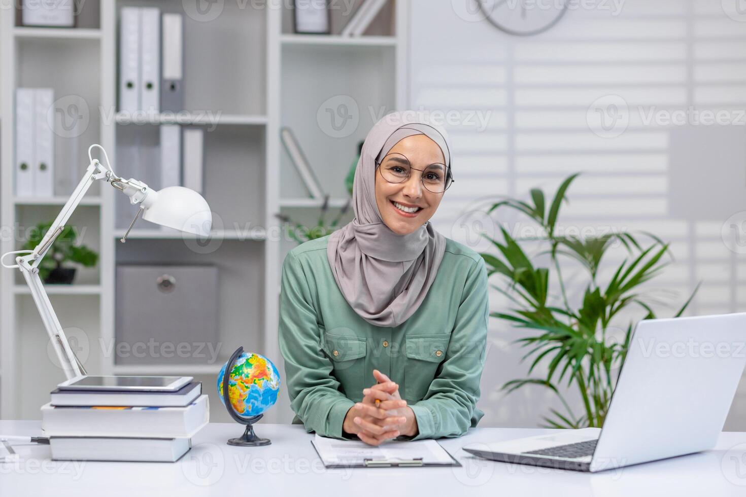 Portrait of happy teacher, woman in hijab sitting at desk smiling and looking at camera, muslim woman working remotely, teaching students and pupils using laptop in home office. photo