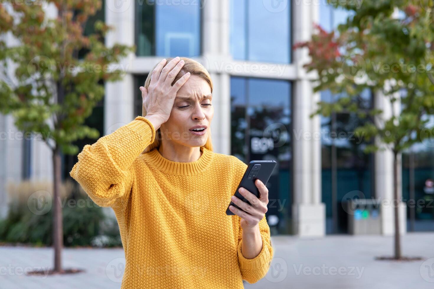 A middle-aged woman appears distressed and worried while she holds a smartphone on a sunny day with an urban background. Her expression conveys shock and confusion. photo