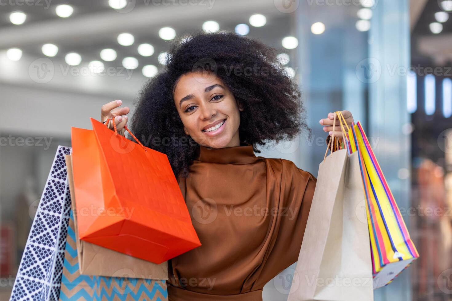 retrato de contento satisfecho compras mujer, africano americano mujer con Rizado pelo dentro supermercado sonriente y mirando a cámara, participación pantalones en manos. foto