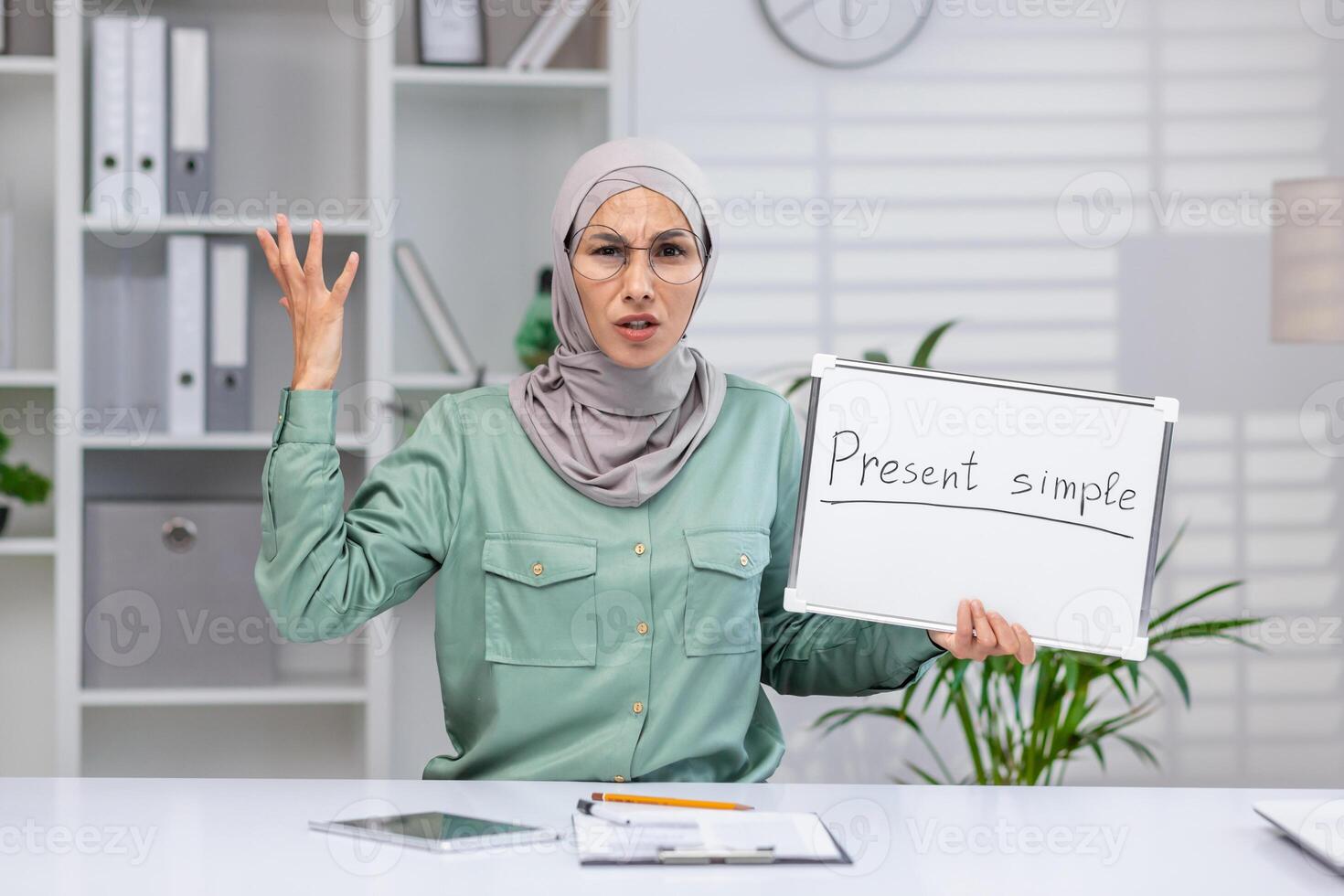 Teacher explaining present simple tense, holding whiteboard in classroom, expressing frustration or confusion, in a modern educational environment. photo